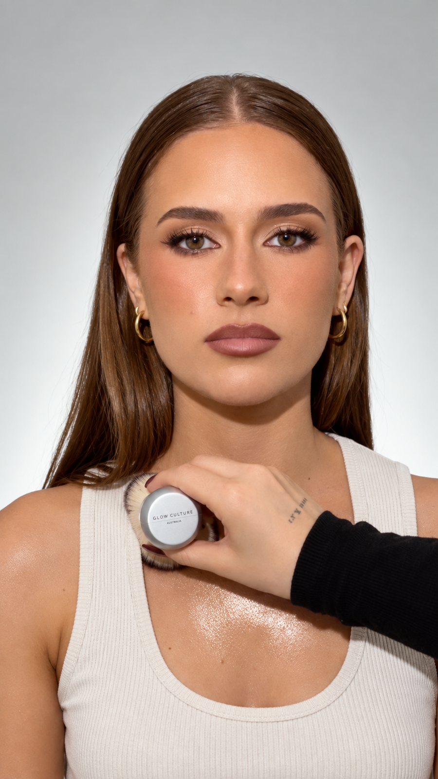 A close-up showing The Glow Brush being used to apply and buff a glossy, hydrating body oil or bronzer onto a woman's arm