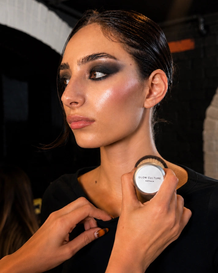 A close-up shot of a model with glowing, dewy skin holding a dense, glow brush up to her face